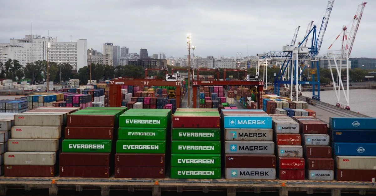 Vista aérea de coloridos contenedores de transporte en un puerto bullicioso con grúas y el perfil urbano de la ciudad.