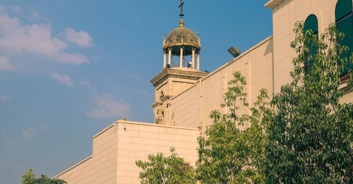 Una torre de iglesia histórica con cúpula en Damasco, rodeada de vegetación bajo un cielo despejado.