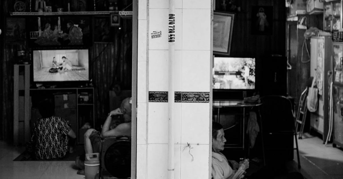 Photographie en noir et blanc de personnes à l'intérieur d'une maison vietnamienne, regardant la télévision, illustrant la vie quotidienne.