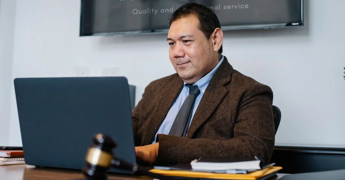 Un abogado trabajando en una laptop en una oficina legal, vestido con traje y corbata, con un mazo sobre el escritorio.